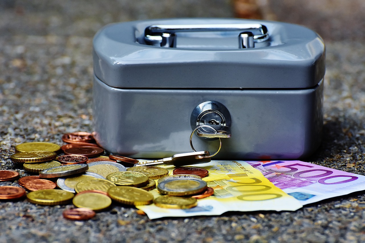A locked gray cash box with a key, surrounded by Euro coins and banknotes, symbolizing savings and financial security.
